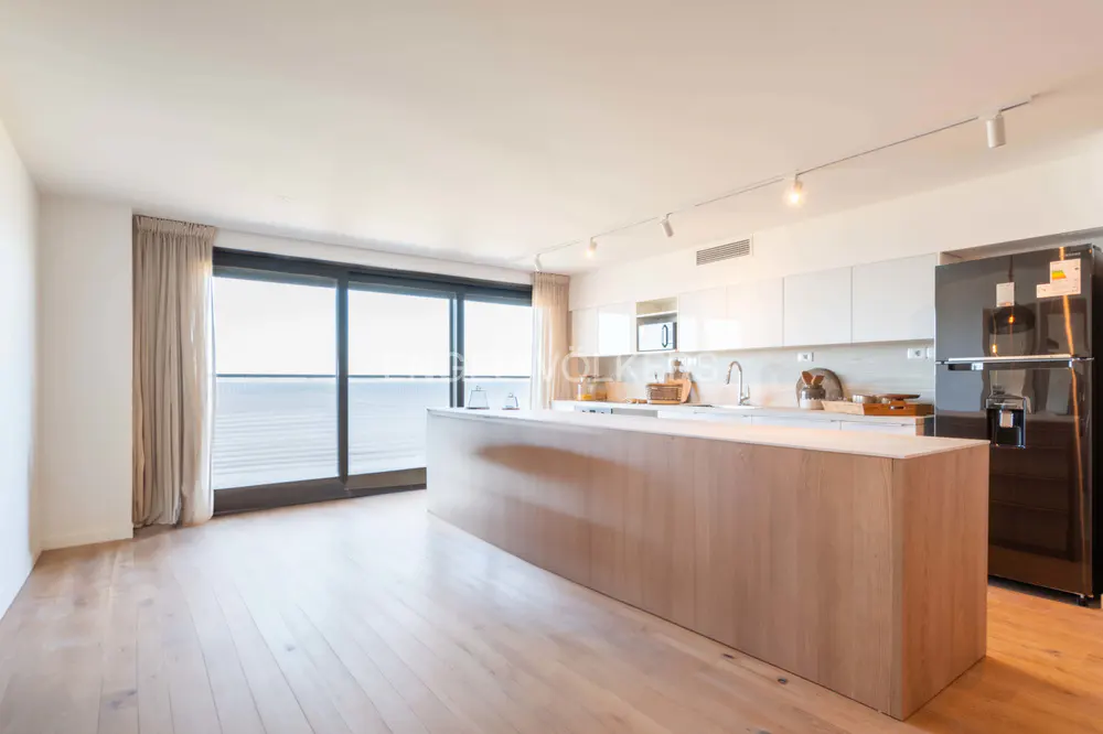 Bright, modern kitchen with wood floors, a large island, white cabinets, and a black refrigerator. A large window overlooks the ocean.