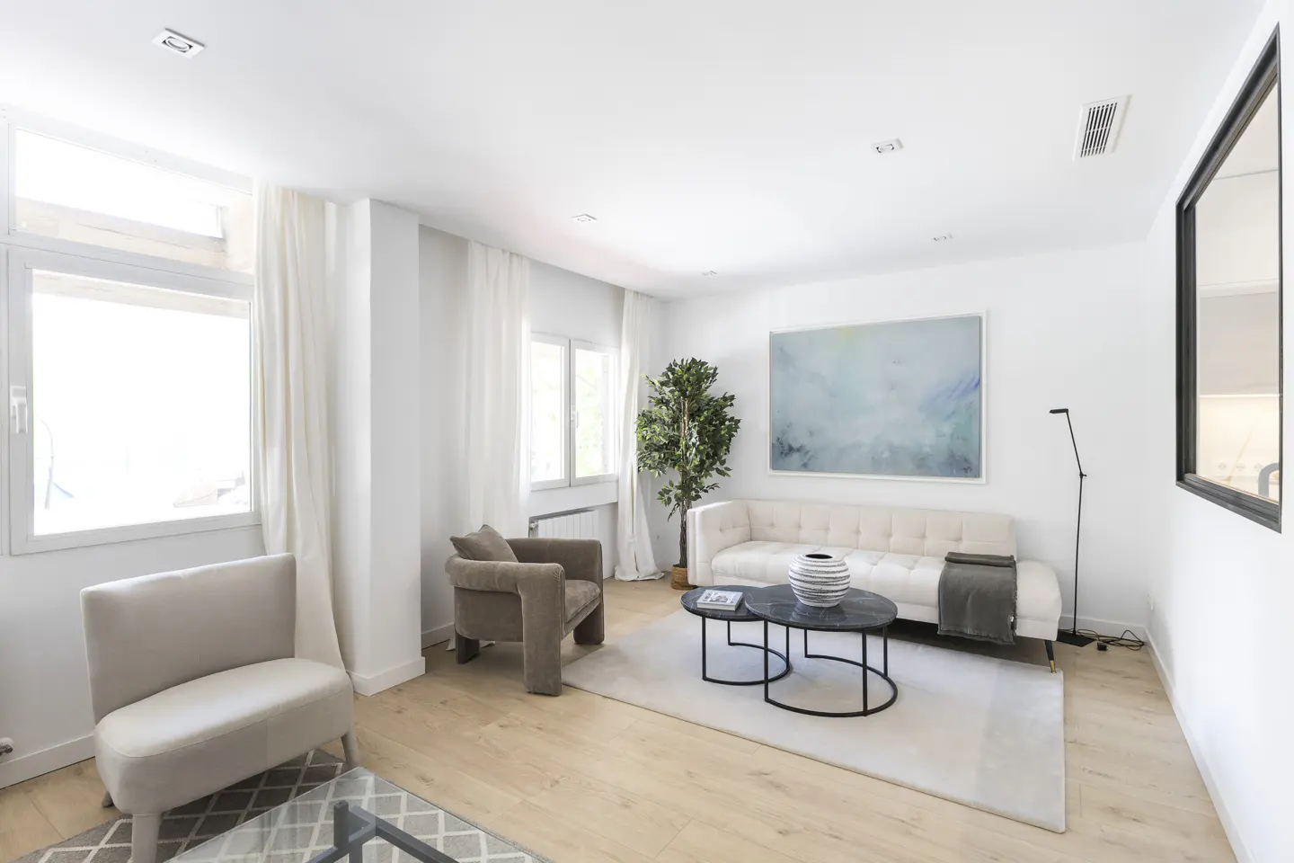 Bright living room with white walls, light wood floors, and neutral furniture. A blue abstract painting hangs above a cream sofa.