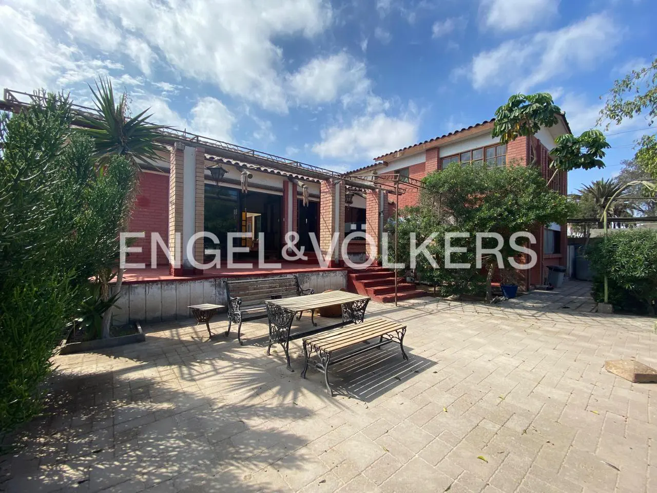 Exterior view of a red brick house with a patio, featuring a table, benches, and lush greenery under a partly cloudy sky.