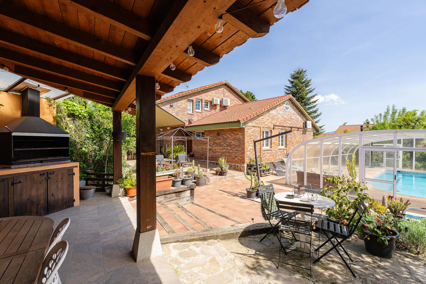 Outdoor patio with a wooden pergola, grill, table, chairs, and a pool with a white enclosure in the background.