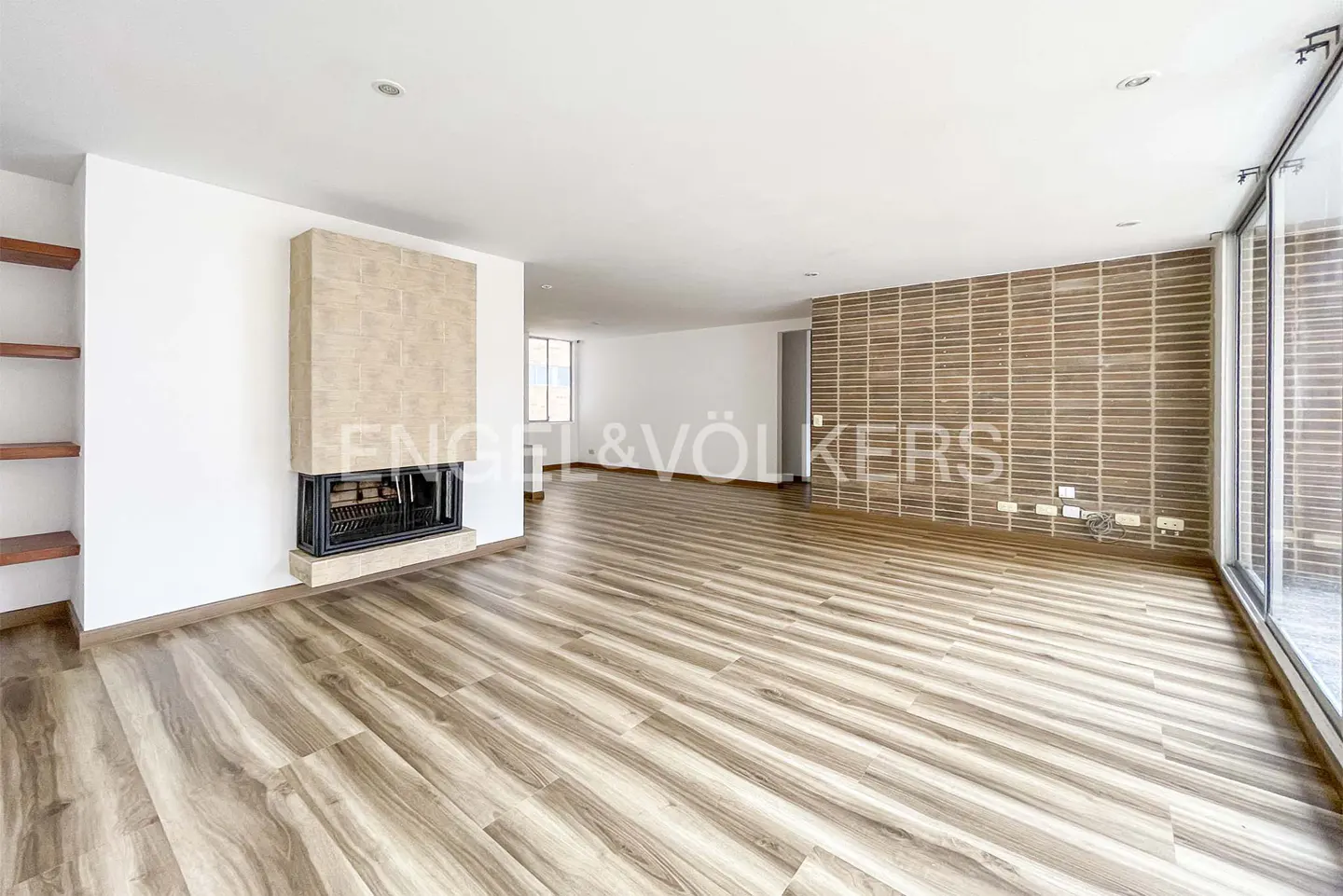 A bright, empty living room with wood floors, a brick accent wall, and a fireplace with a stone surround.