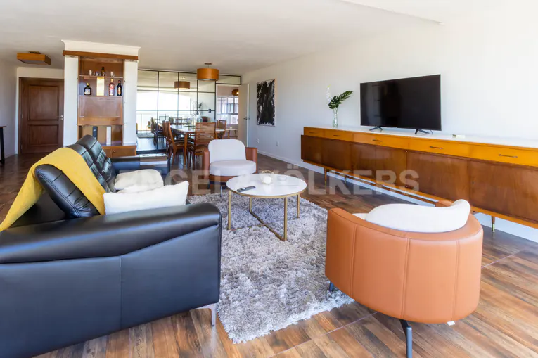 Bright living room with black leather sofa, brown chairs, and a dining area visible through a glass partition. A TV sits atop a wooden cabinet.