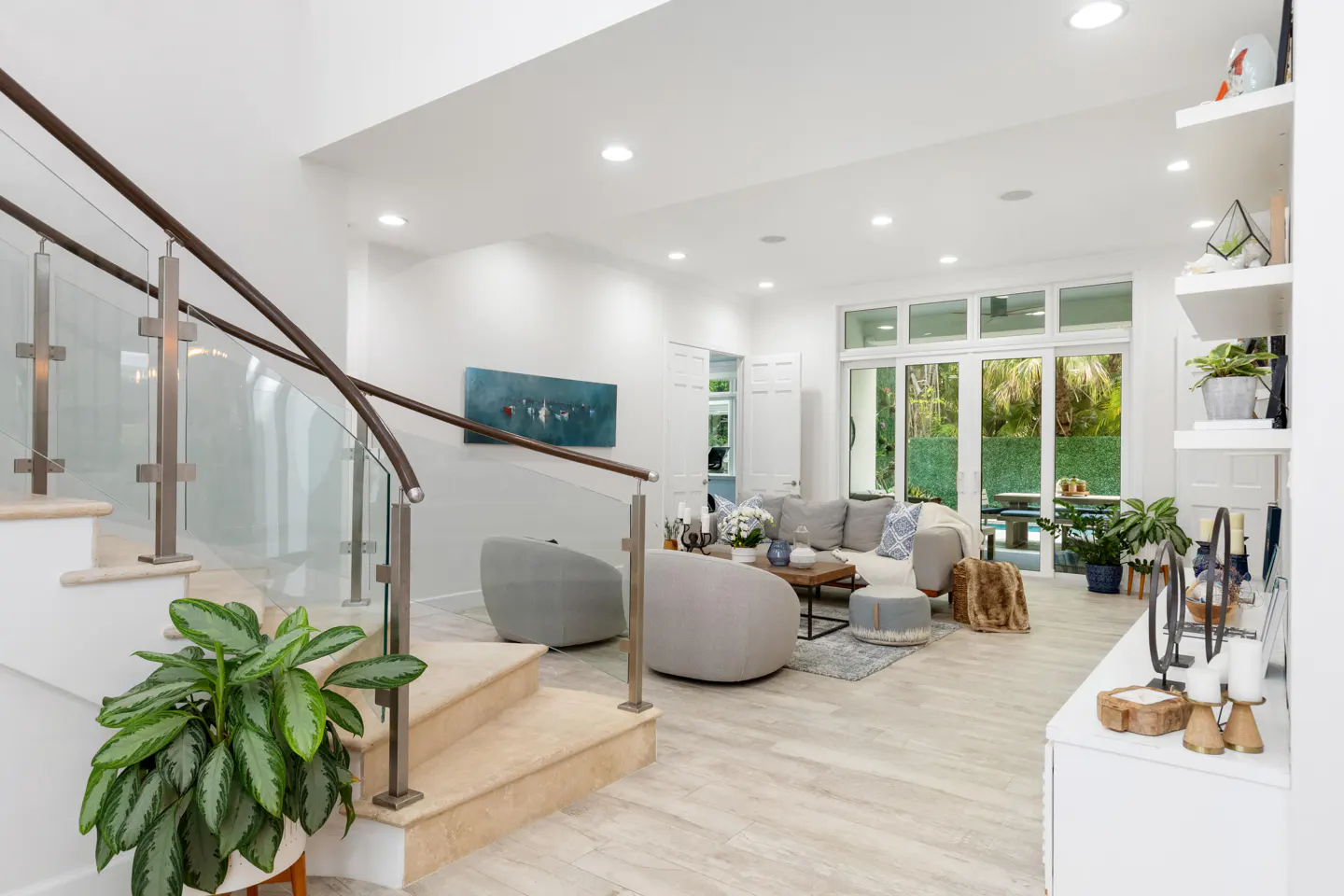 Bright, modern living room with staircase. Beige tile floors, white walls, and glass railing. Gray chairs and sofa face sliding glass doors to the patio.