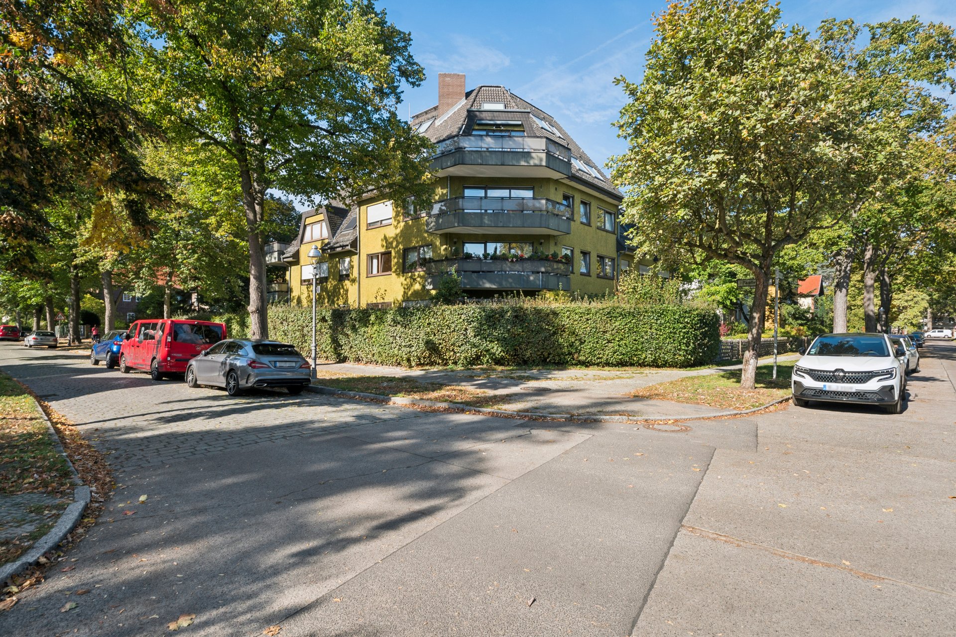 A yellow multi-story apartment building with balconies, surrounded by trees and a hedge, on a sunny street with parked cars. [AI-generated text]