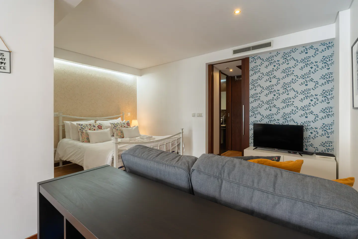 Studio apartment with a gray sofa, TV, and a white metal bed frame with white bedding and floral pillows.