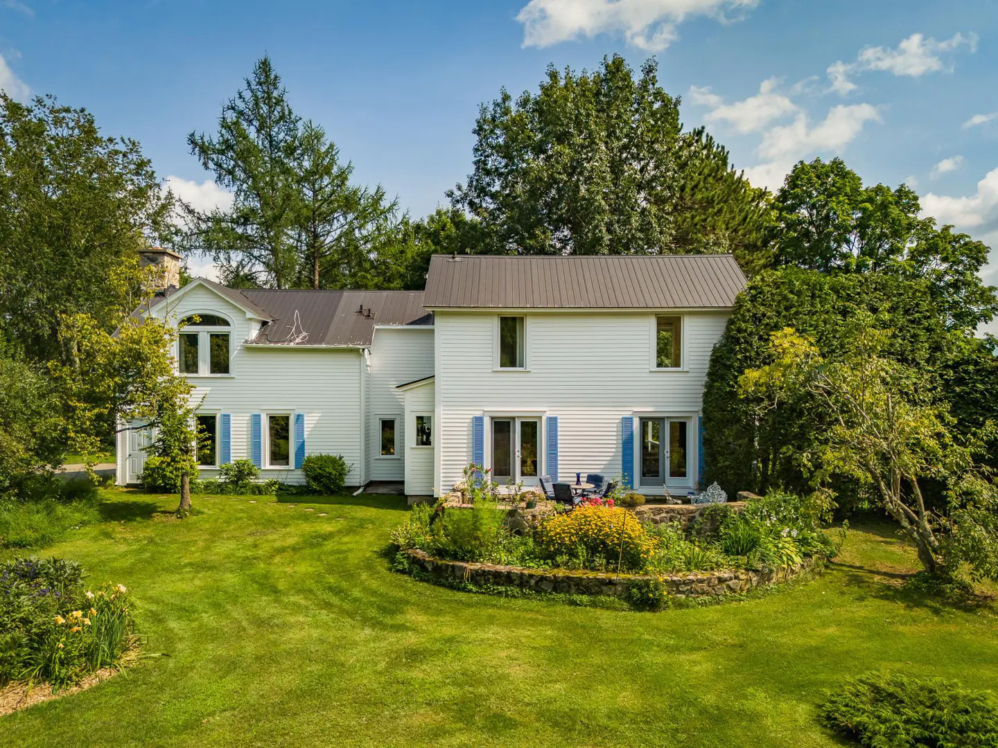 Two-story white farmhouse with blue shutters, a gray roof, and a green lawn on a sunny day.