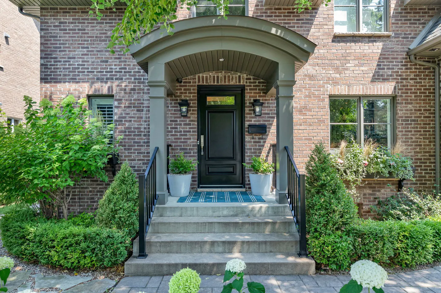 Brick house facade with a black front door under a gray arched porch. Steps lead up with black railings, flanked by green bushes and potted plants.