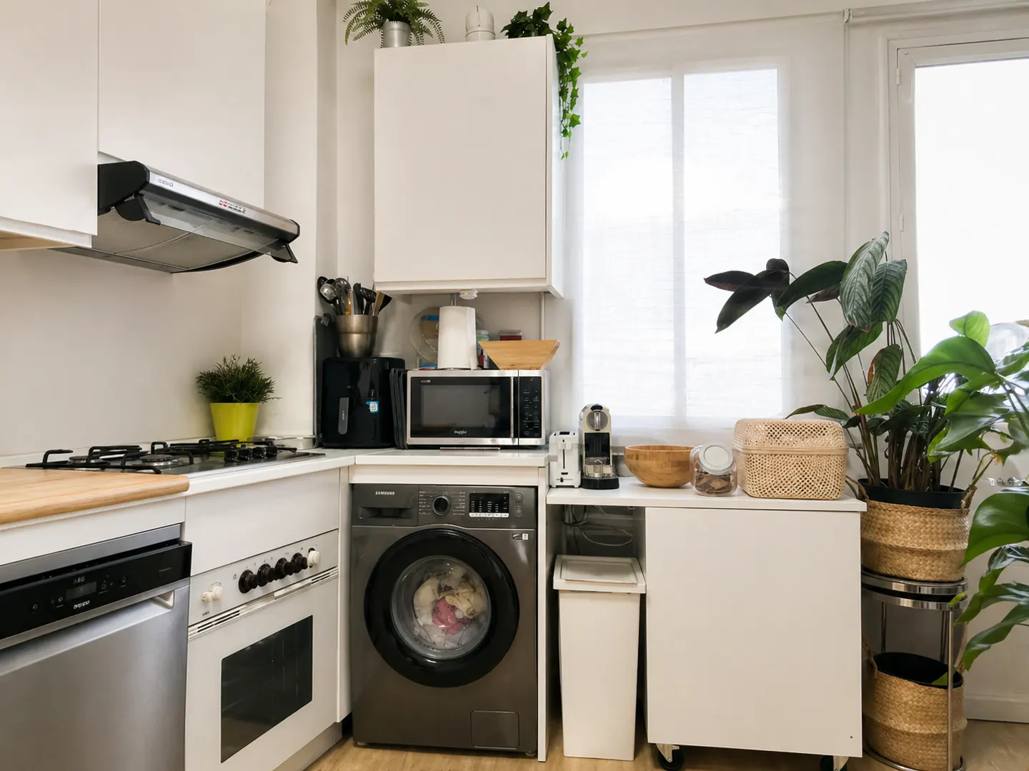 Bright kitchen with white cabinets, stainless steel appliances, and a gray washing machine. Potted plants add greenery near a window.
