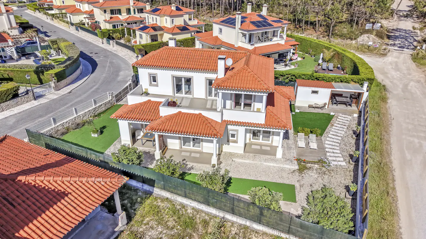 Aerial view of a white two-story house with an orange tile roof, surrounded by green lawns and trees in a residential area.