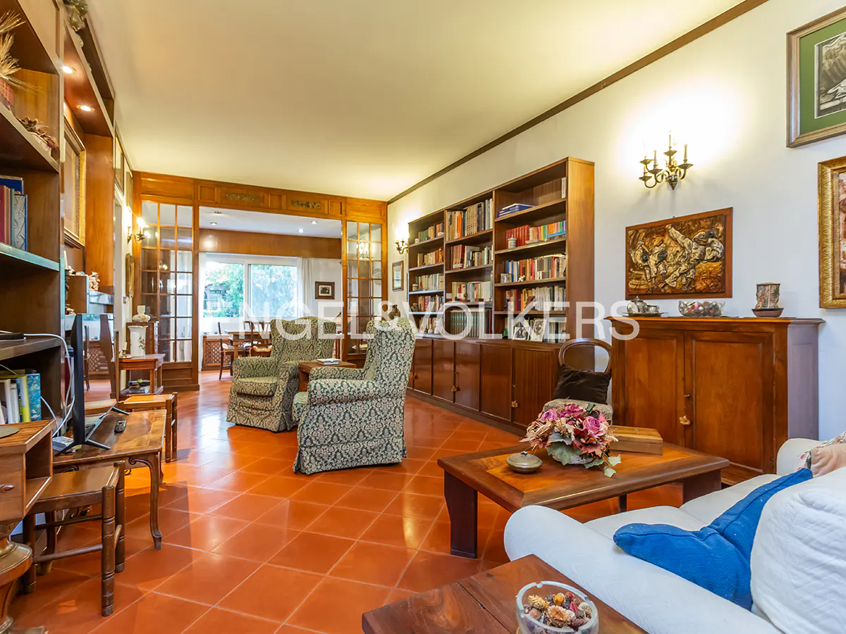 A living room with terracotta tile floors, bookshelves, and patterned armchairs. A white sofa with blue pillows is in the foreground.