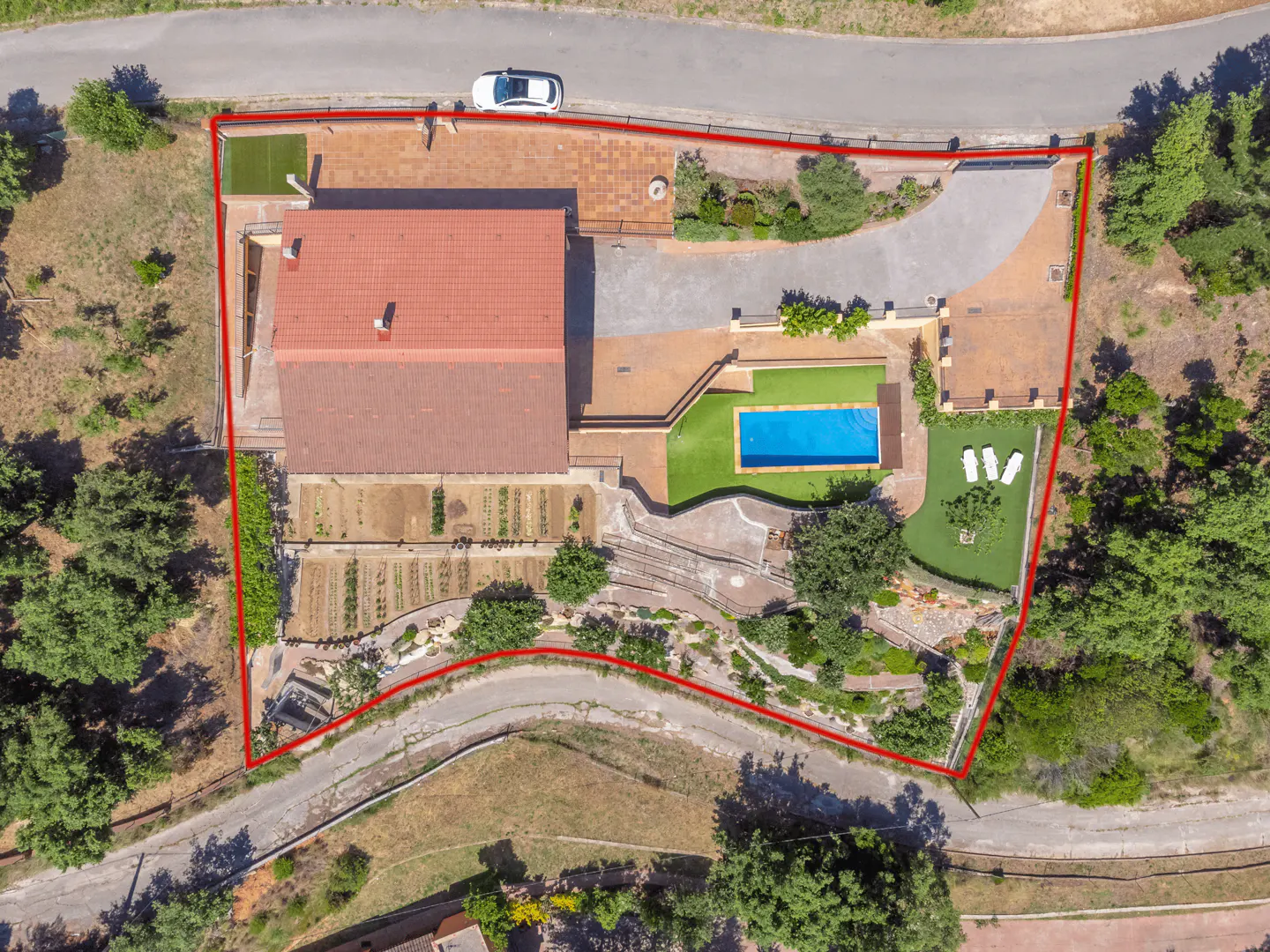 Aerial view of a property with a red tile roof, pool, garden, and a red boundary line. A white car is parked near the house.