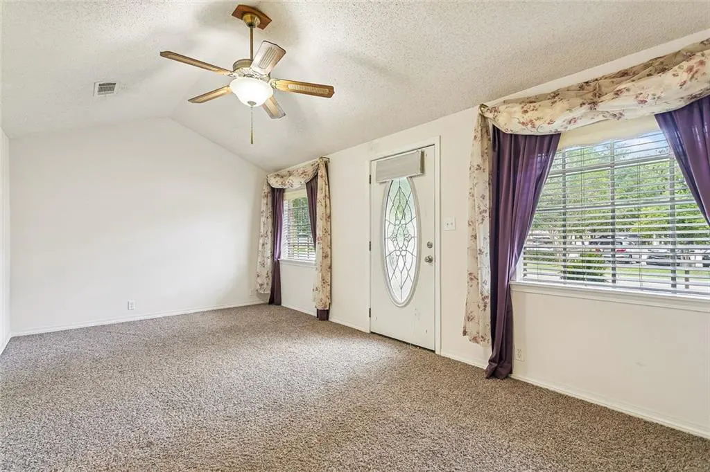 A bright living room with beige carpet, white walls, and a ceiling fan. Windows with purple curtains flank a white front door.