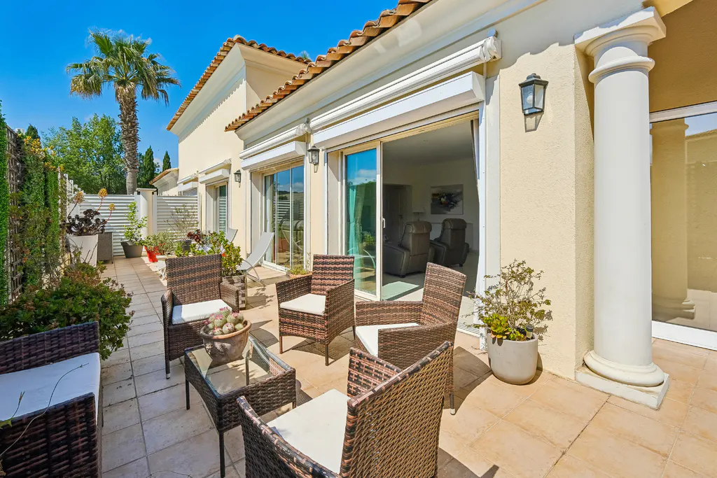 Outdoor patio with wicker furniture, potted plants, and a palm tree under a blue sky. Sliding glass doors lead into the home.