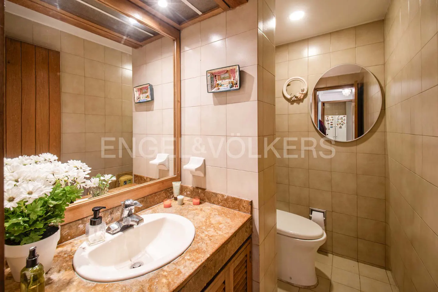 Bathroom with beige tile walls, a marble countertop, and a white sink. A round mirror hangs above the toilet. A vase of white flowers sits on the counter.