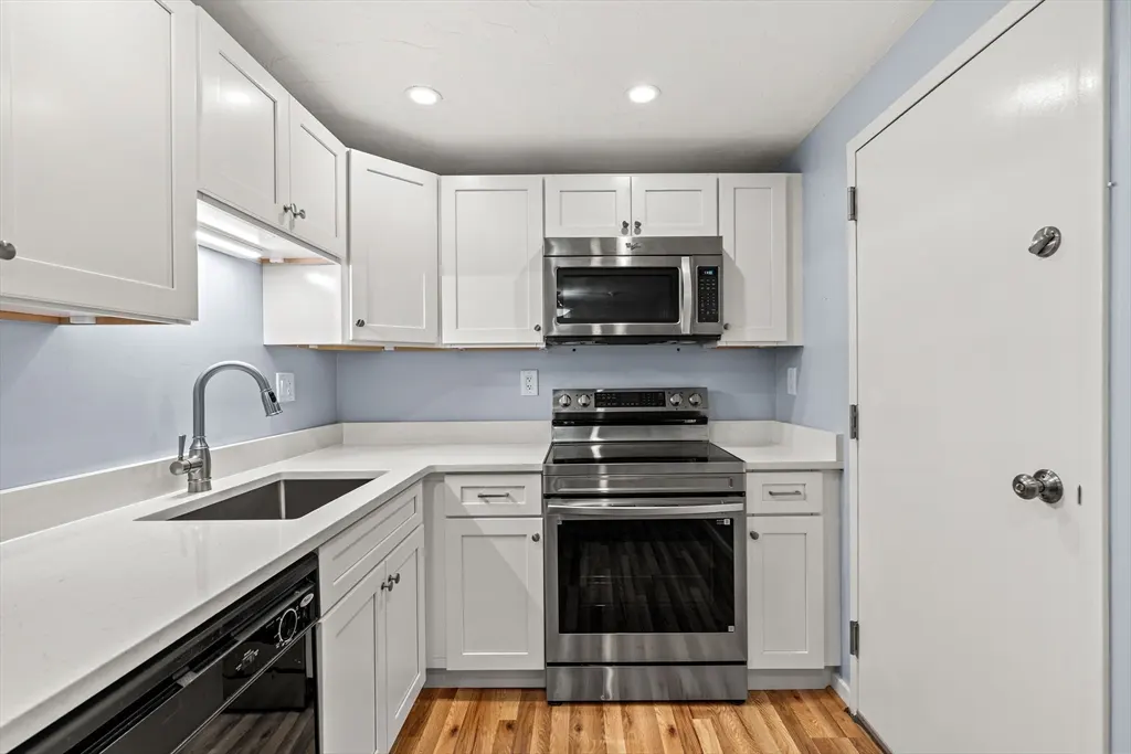 A compact kitchen with white cabinets, stainless steel appliances, light countertops, and wood floors. A white door is on the right.