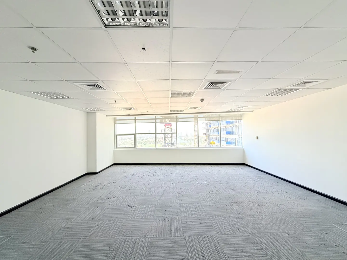 Empty office space with white walls, grey carpet tiles, a drop ceiling, and large windows overlooking a city view.
