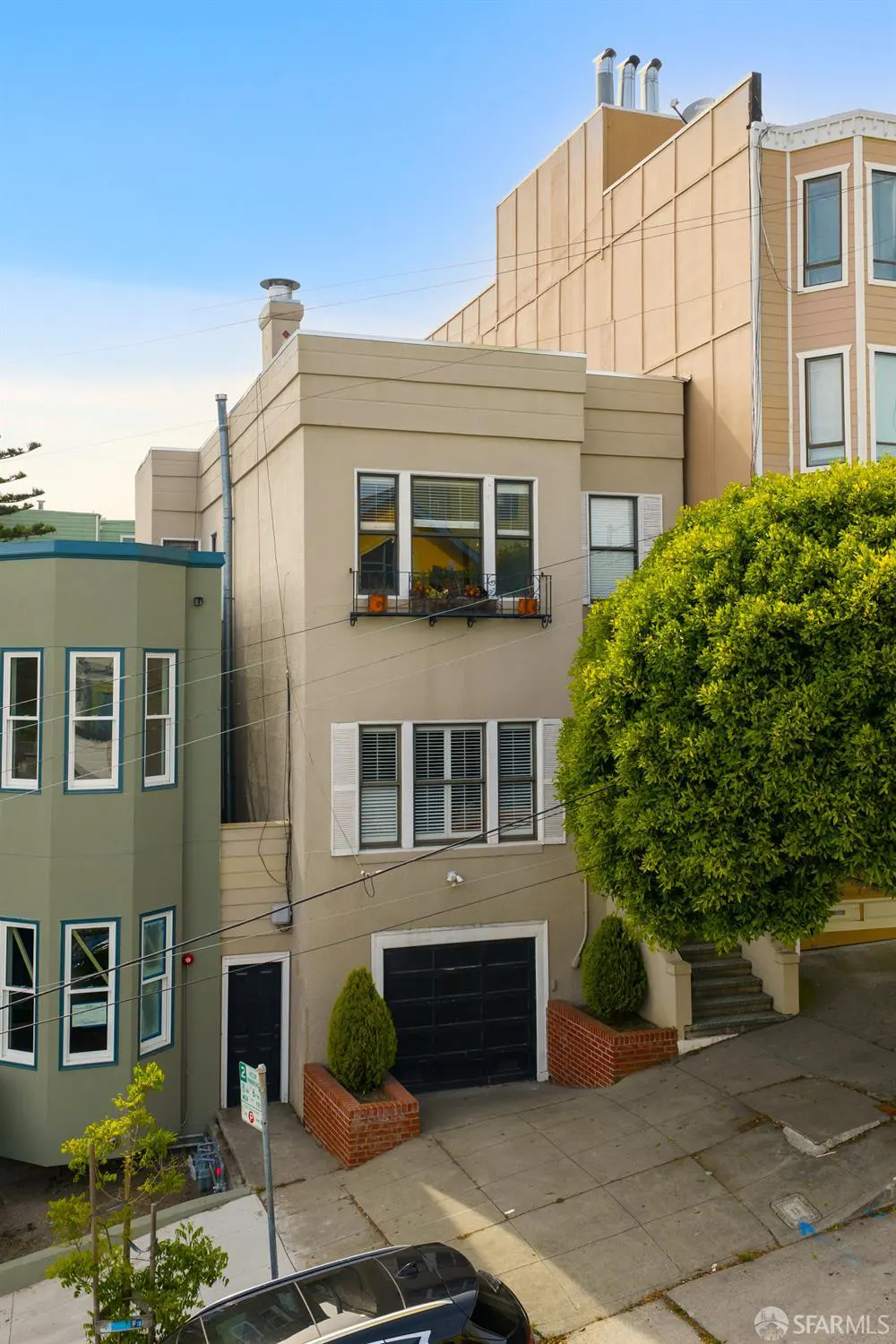 Tan two-story house with a black garage door and white window shutters. Green bushes and trees surround the house. Blue sky.