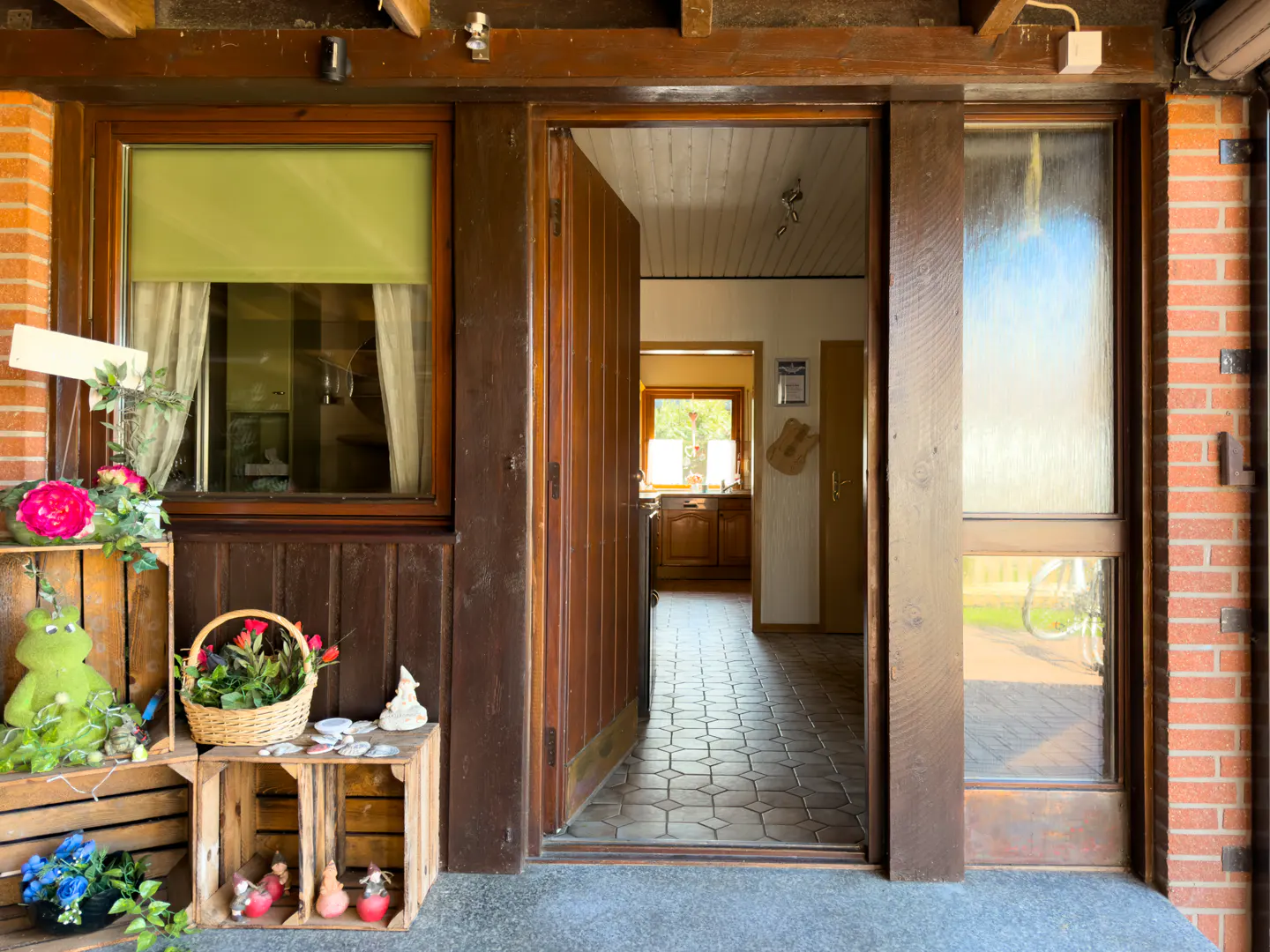 Open wooden front door of a house with brick and wood exterior. Interior view of tiled hallway and kitchen. Crates with flowers decorate the entrance.