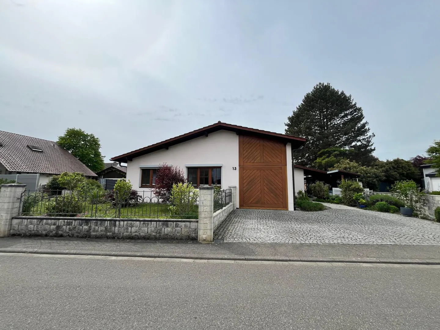 A single-story white house with a brown roof and garage door, number 13, sits behind a stone wall and iron fence.