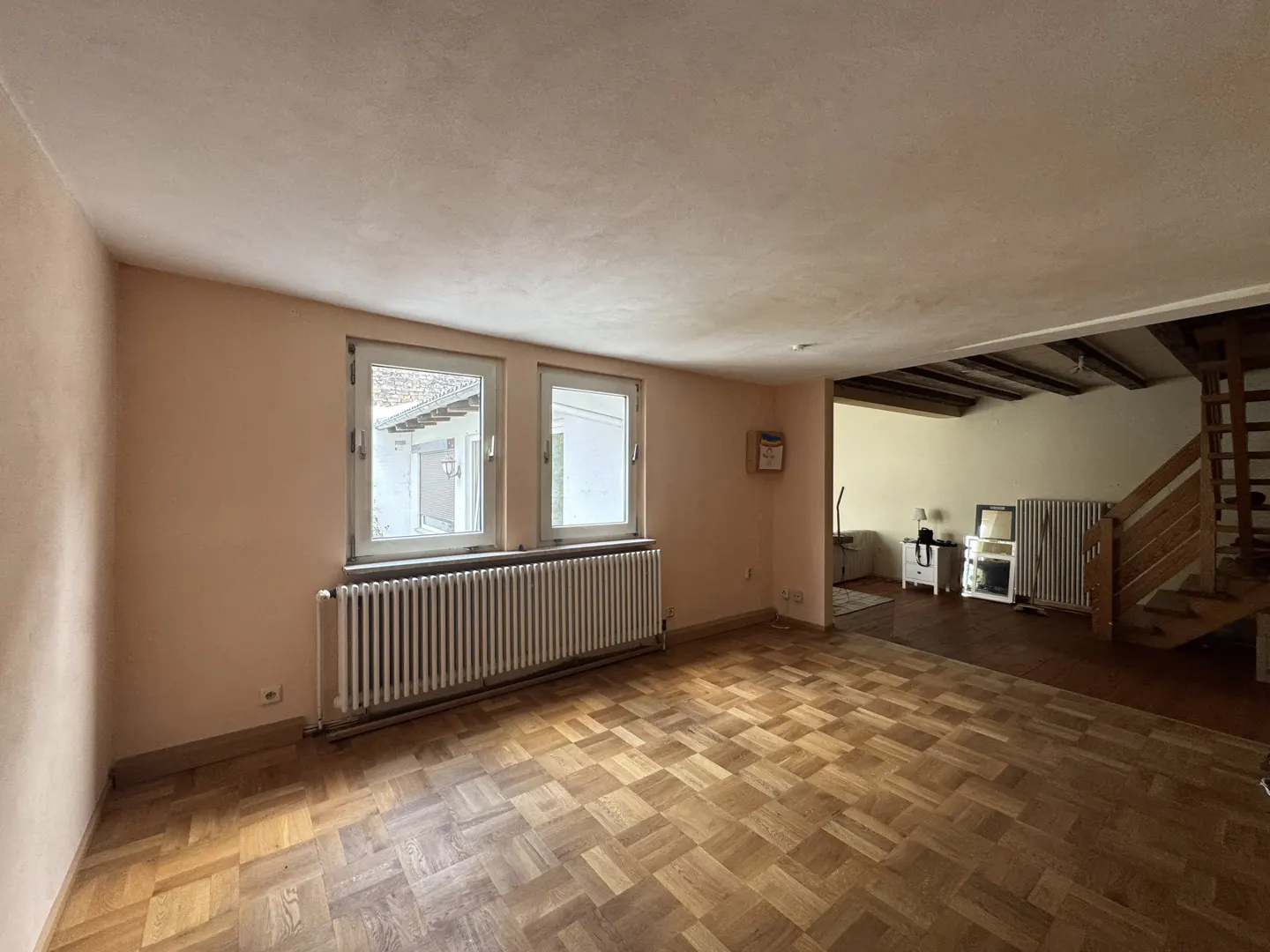 Empty room with light peach walls, parquet flooring, a white radiator under two windows, and a wooden staircase nearby.