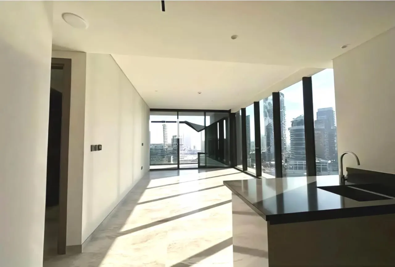 Bright, modern apartment interior with floor-to-ceiling windows offering a city view. A kitchen island with a sink is visible.