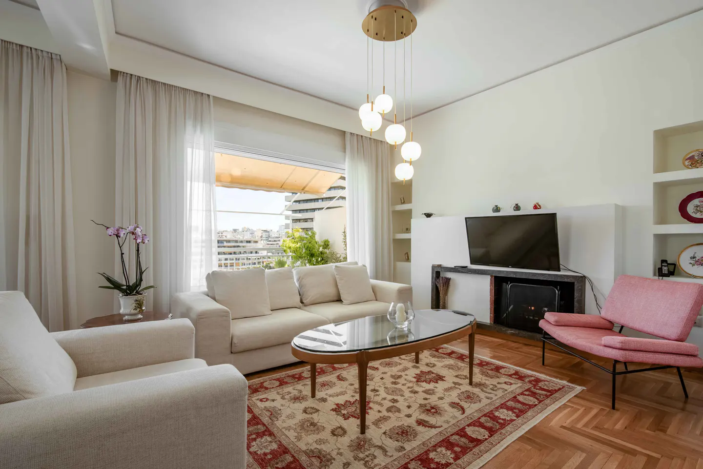 Bright living room with white sofas, pink chair, and glass coffee table on a patterned rug. A city view is visible through the window.