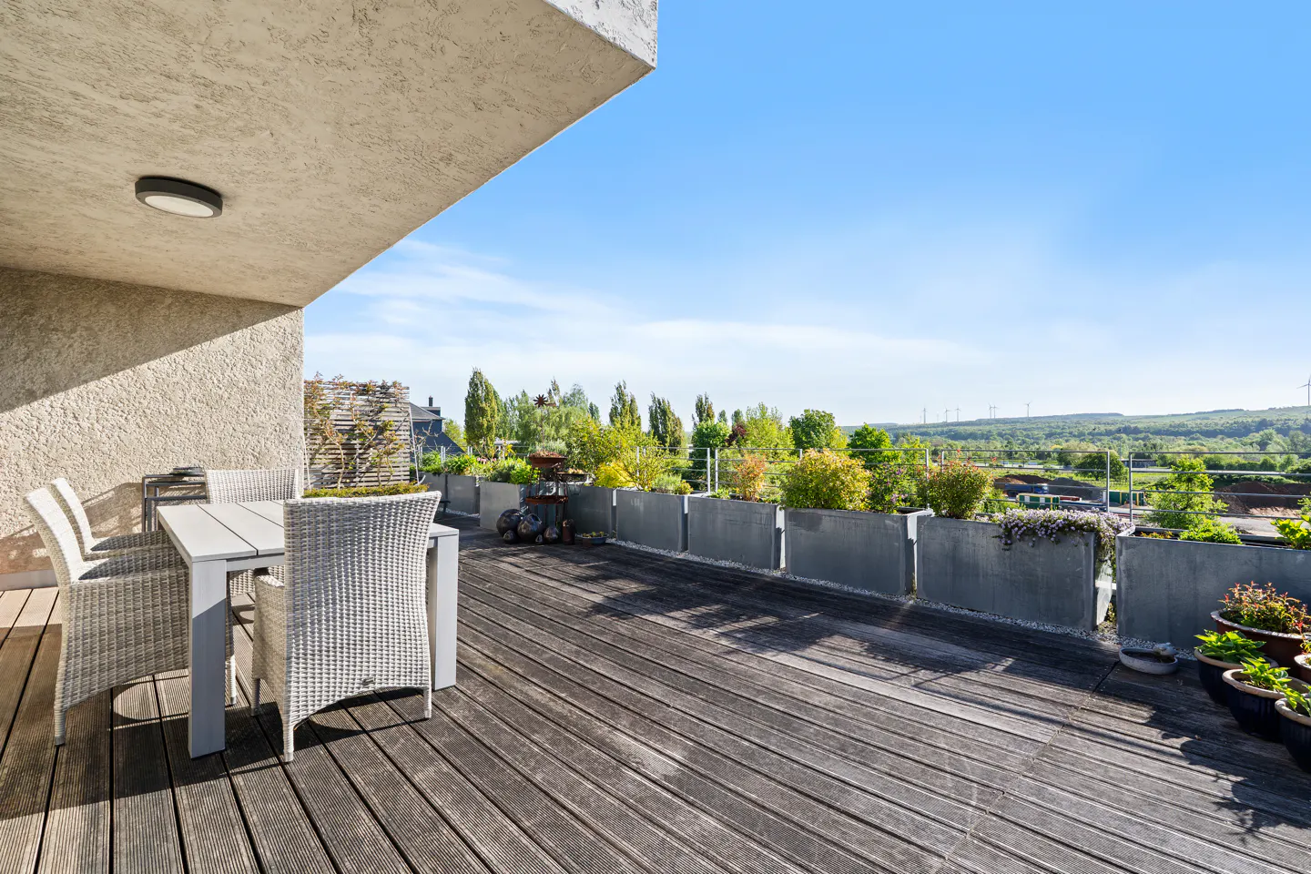 Outdoor patio with wooden deck, table and chairs, and a view of trees and blue sky.