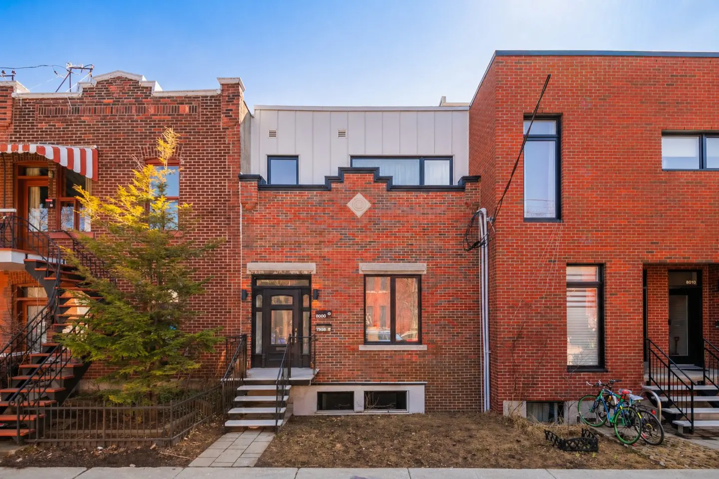 Three brick townhouses stand side-by-side. The center house has a modern white addition on top. A small tree is in front of the left house.