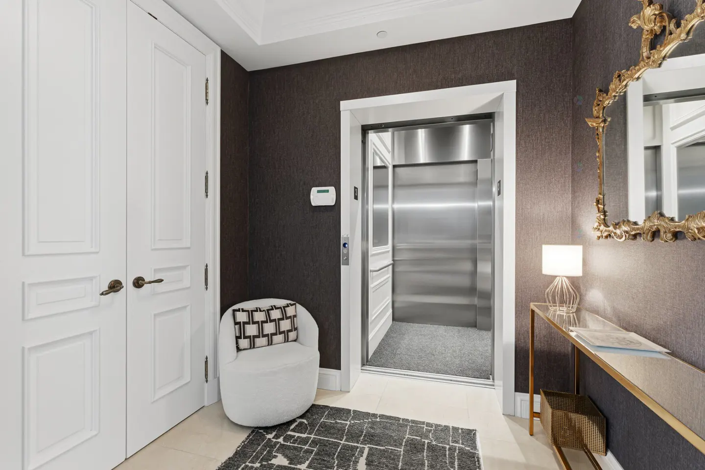 A foyer with dark textured wallpaper, white doors, and a stainless steel elevator. A white chair and gold console table add elegance.