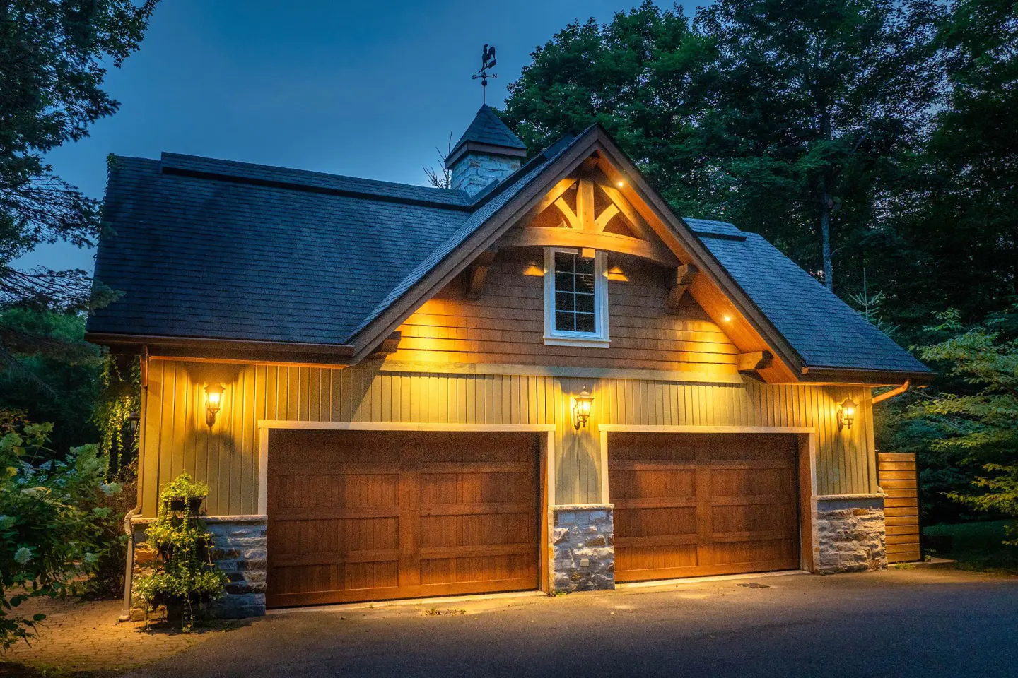 A two-car garage with brown doors, lit by sconces, stands against a dark green forest backdrop.