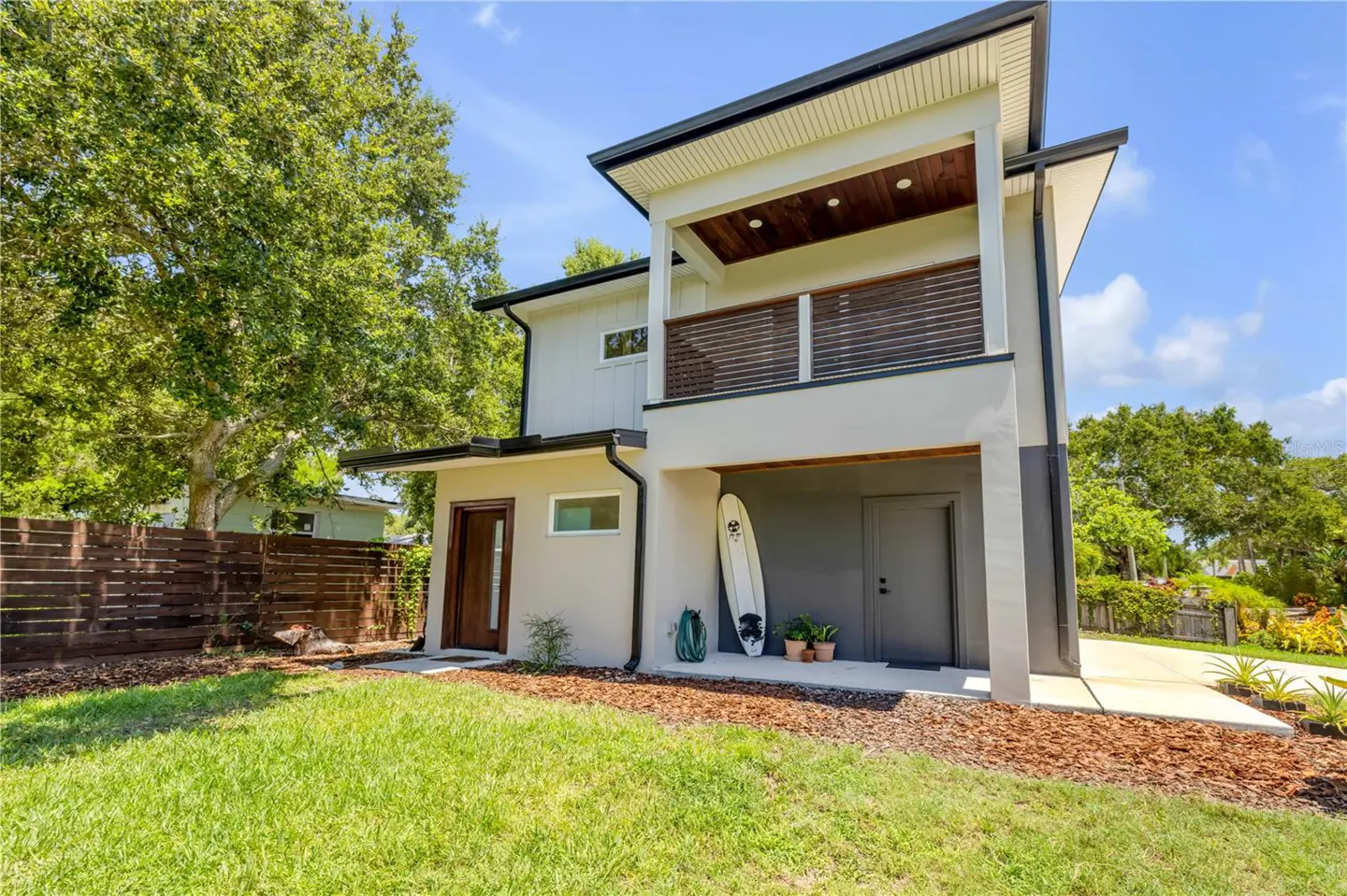 Two-story modern home with a balcony, gray exterior, and a surfboard leaning against the wall. Green lawn and trees surround the property.