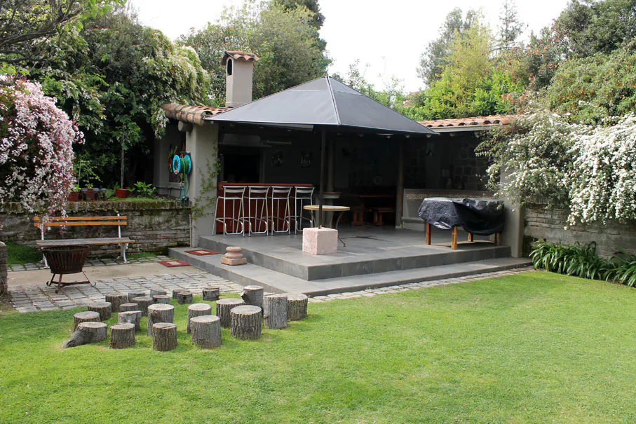 A backyard with a covered patio, bar stools, a grill, and tree stump seating on green grass.