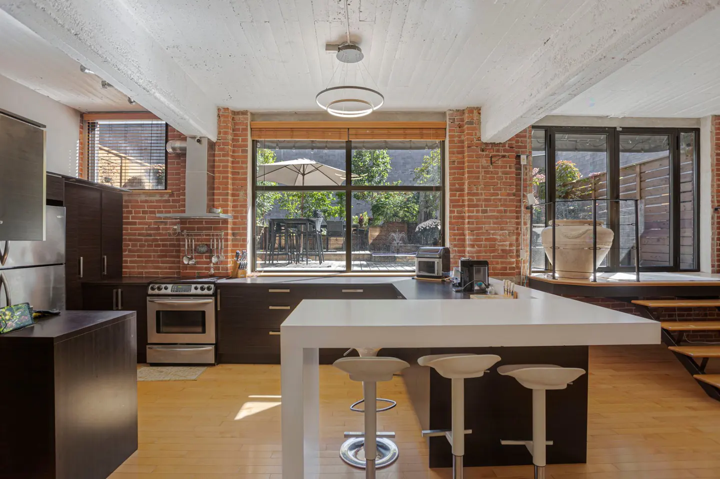 A modern kitchen with brick walls, dark cabinets, stainless steel appliances, and a white island with stools. A large window overlooks a patio.