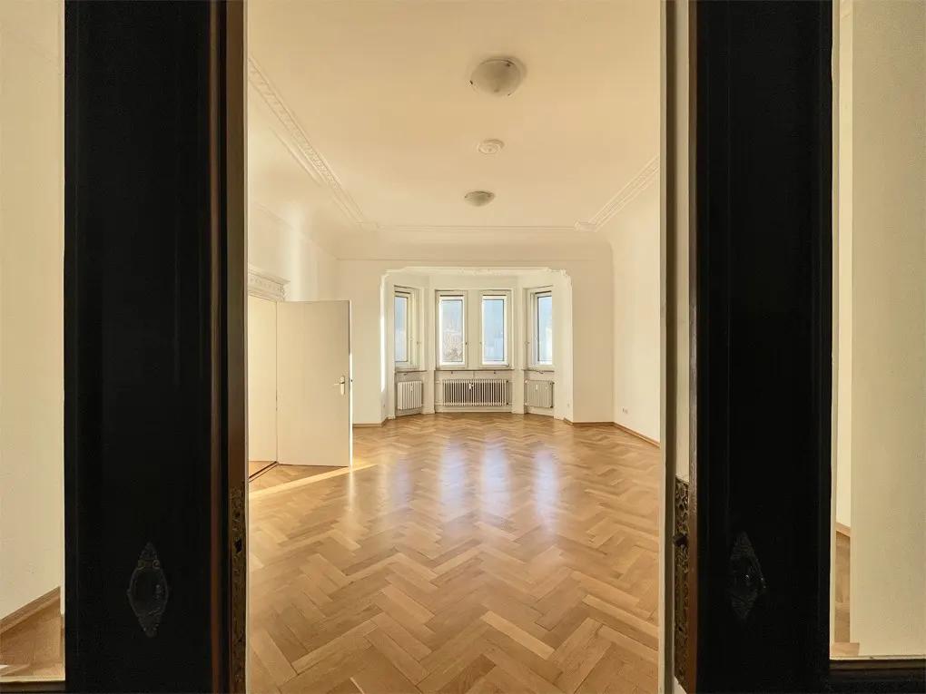 View through dark wood doorway into bright, empty room with herringbone wood floor and bay window.