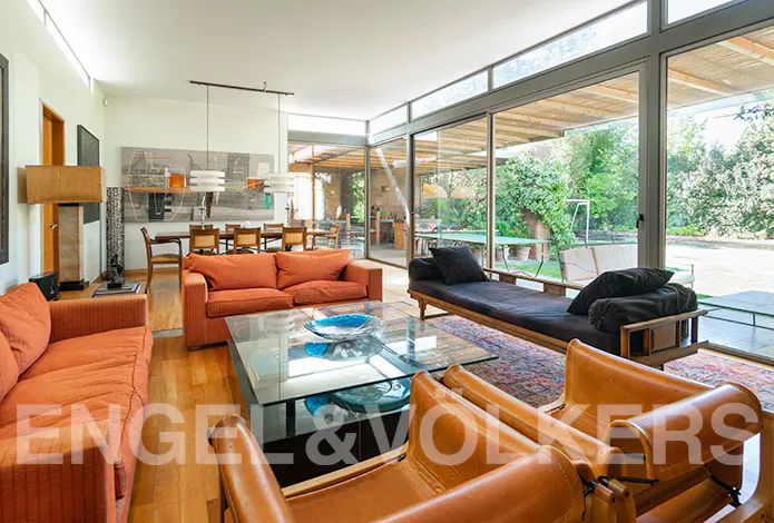 Bright living room with orange sofas, leather chairs, and a glass table. Large windows overlook a green garden.
