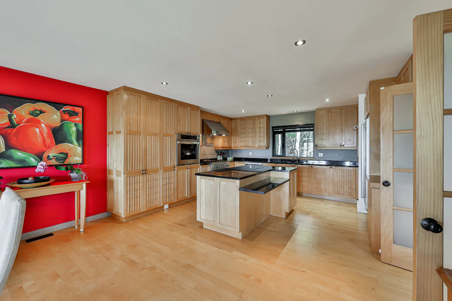 A bright kitchen with light wood cabinets, black countertops, and a central island. A red wall features a pepper painting.