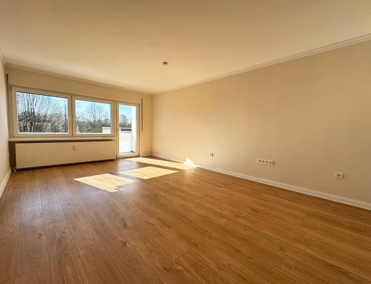 Bright, empty room with wood floors, beige walls, and large windows. Sunlight streams in, highlighting the spaciousness. A radiator sits under the windows.