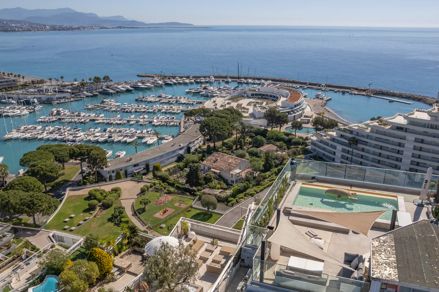 Aerial view of a marina filled with boats, buildings, and a swimming pool on a sunny day.