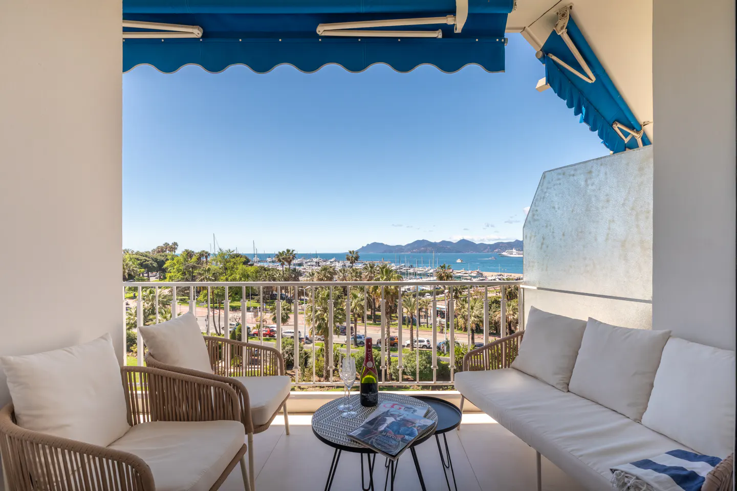 Balcony view with white furniture, blue awning, and a table with wine overlooking a harbor with boats and palm trees.