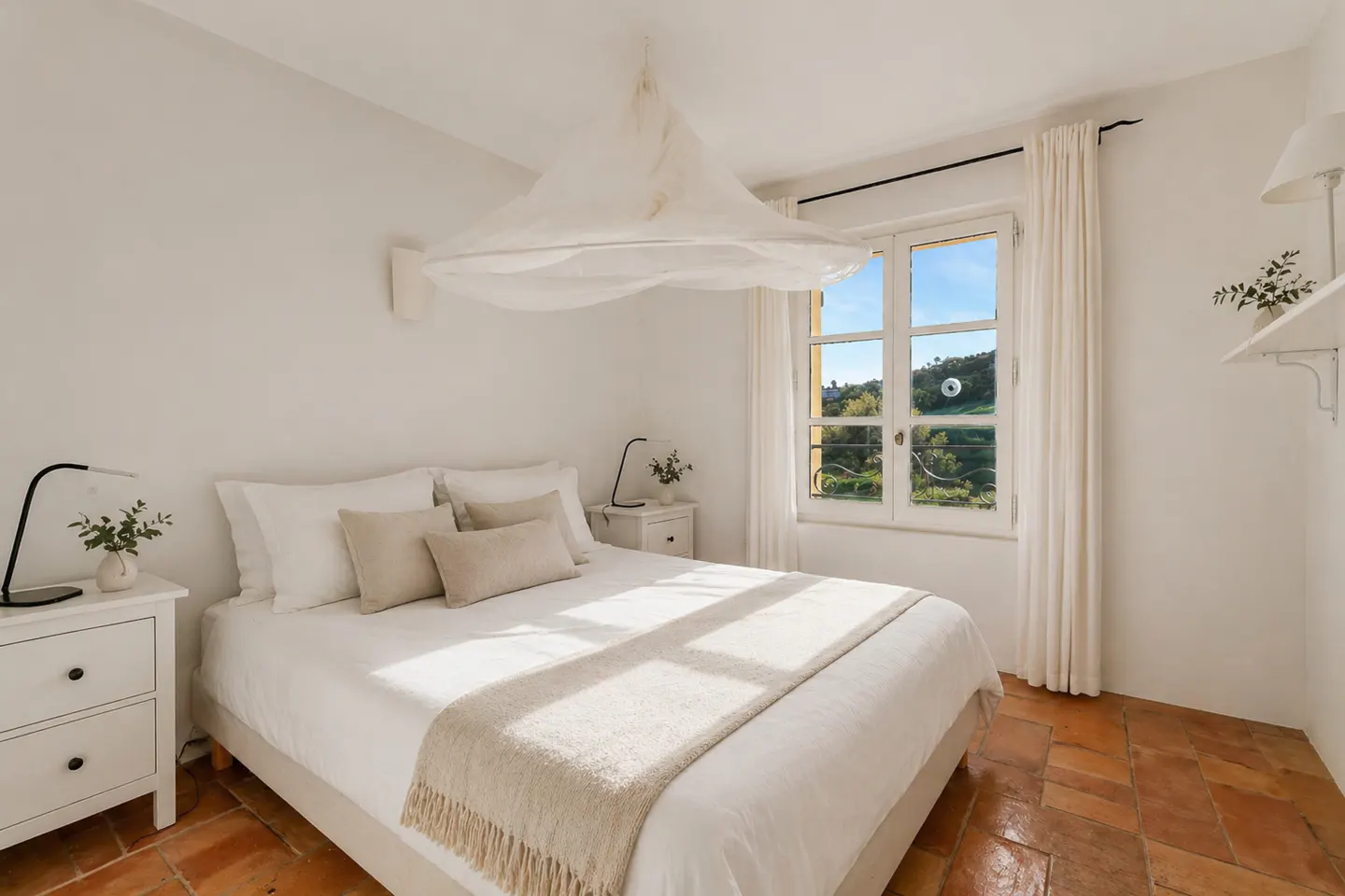 Bright bedroom with white walls, terracotta tile floor, and a large window with a view of green hills. A white bed with a beige throw blanket is centered.