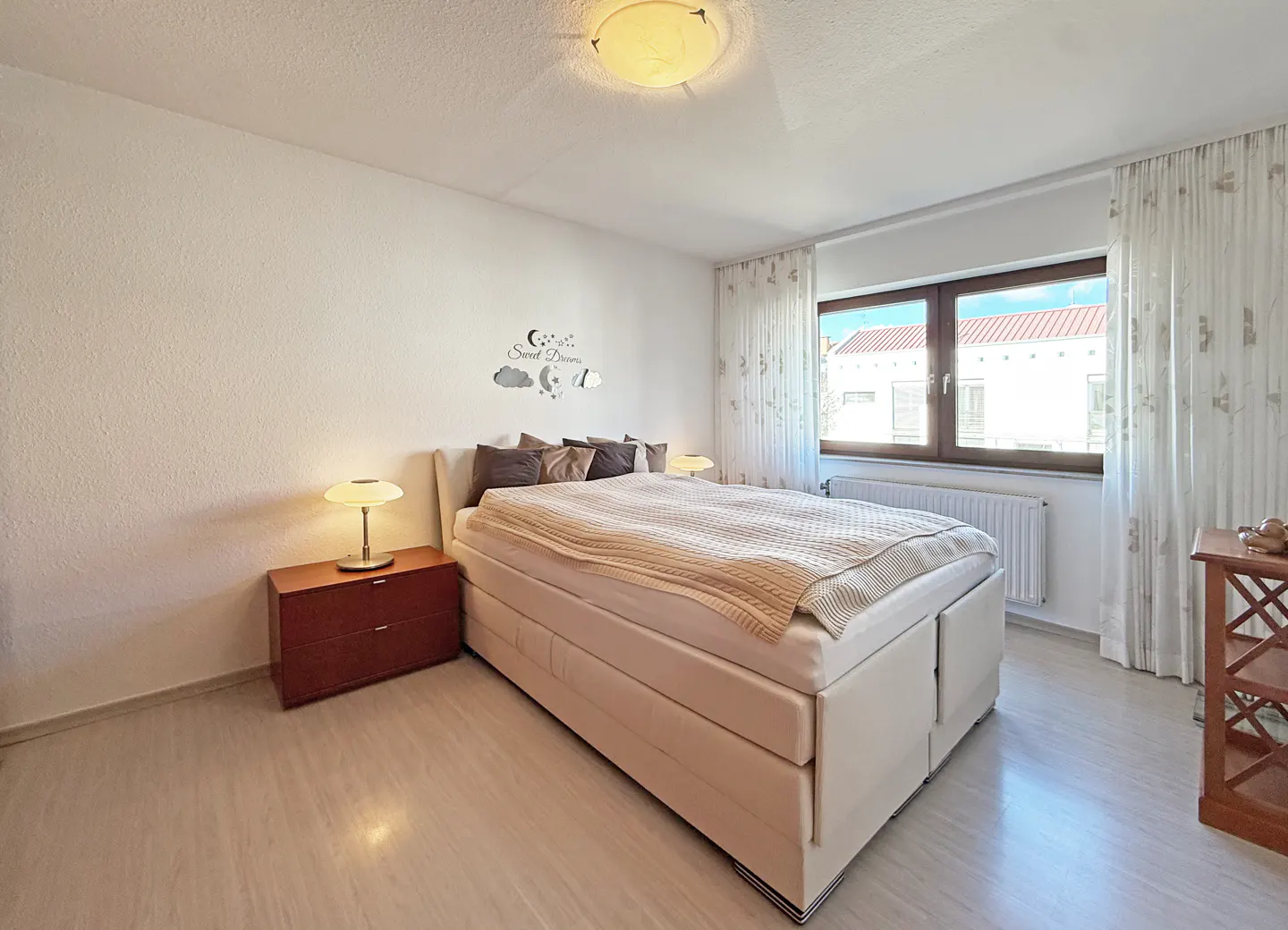 Bright bedroom with a beige bed, brown pillows, and a light beige blanket. A brown nightstand with a lamp is on the left. A window with white curtains is on the right.
