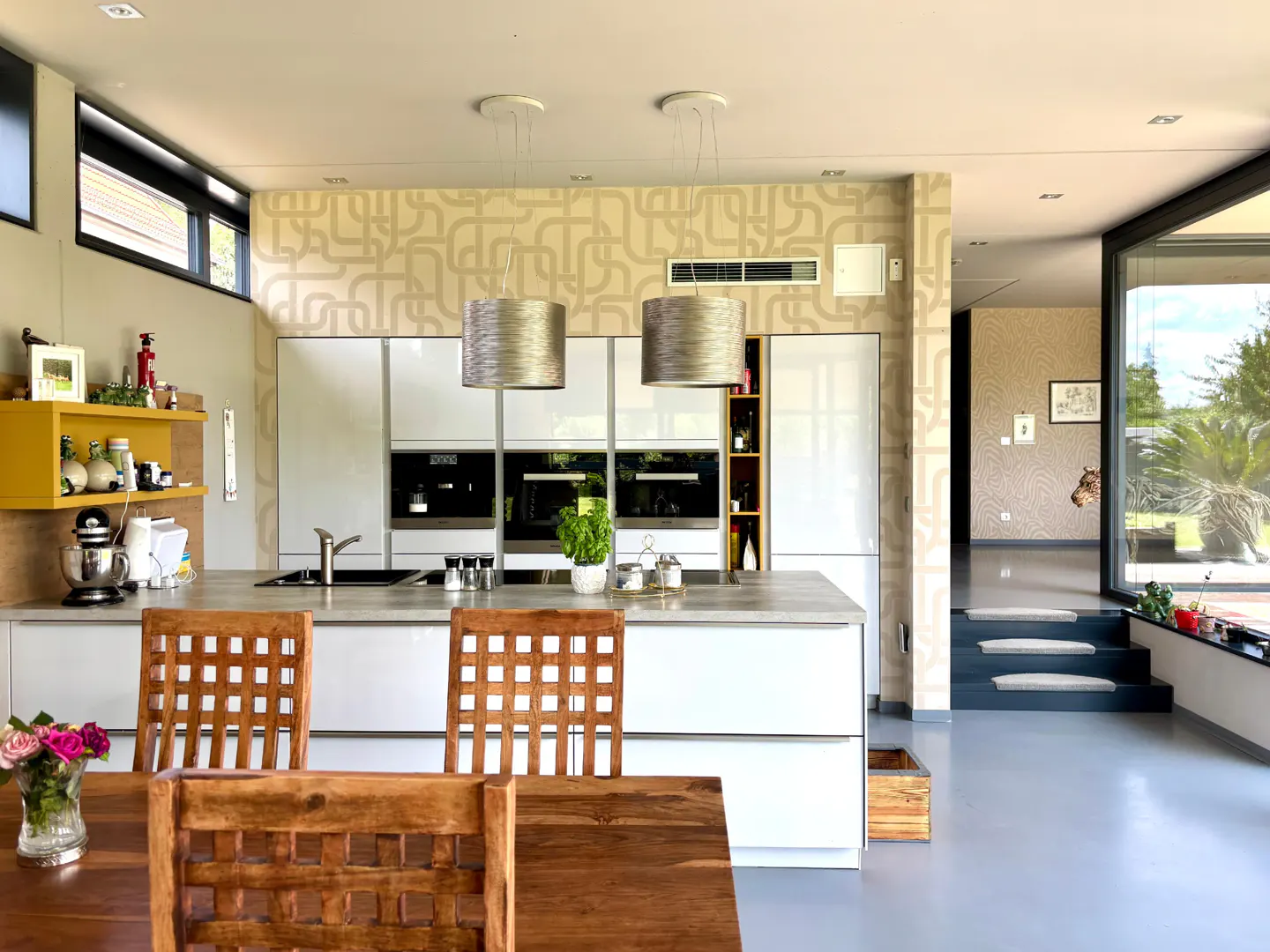 A modern kitchen with white cabinets, a gray countertop island, and wooden chairs at a dining table. Two silver pendant lights hang above the island.