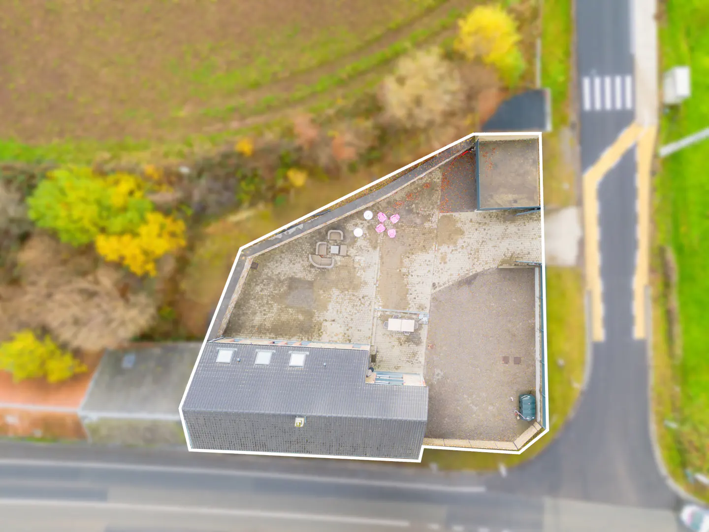 Aerial view of a building with a gray roof and a large, flat rooftop patio with outdoor furniture. A road and green fields surround the property.