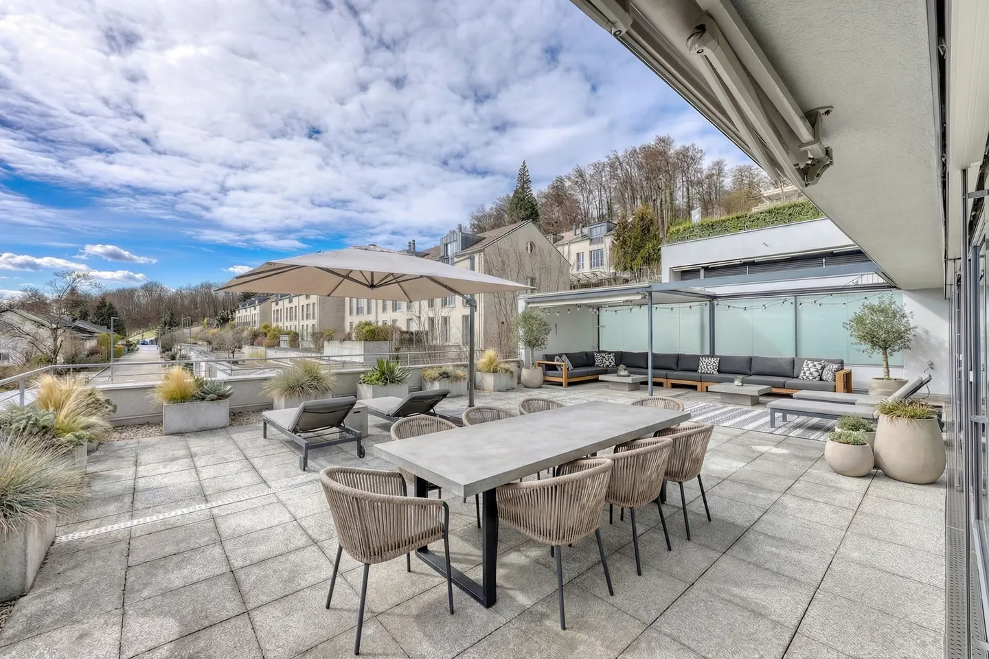 A rooftop patio with a large table, chairs, lounge chairs, and a covered seating area. The patio is surrounded by plants and overlooks a street.