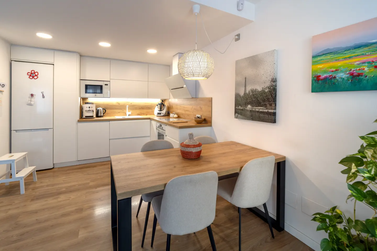 Bright, modern kitchen with white cabinets, wood countertops, and a dining table with four chairs. Artwork adorns the white walls.