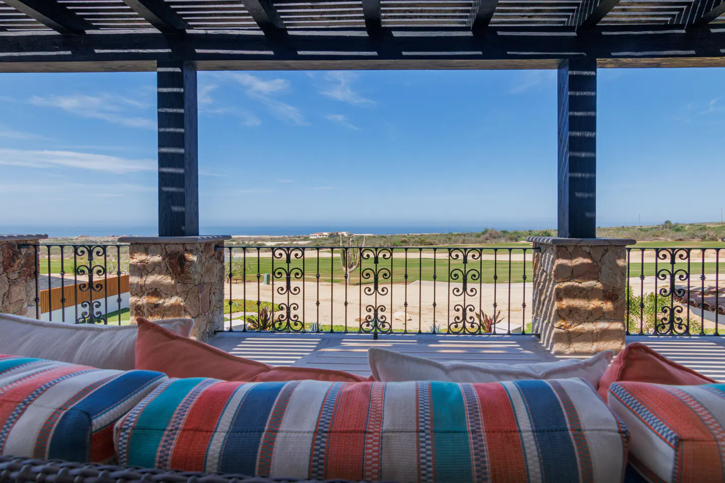 View from a shaded patio with striped cushions, looking out over a wrought-iron railing toward a golf course and the ocean.