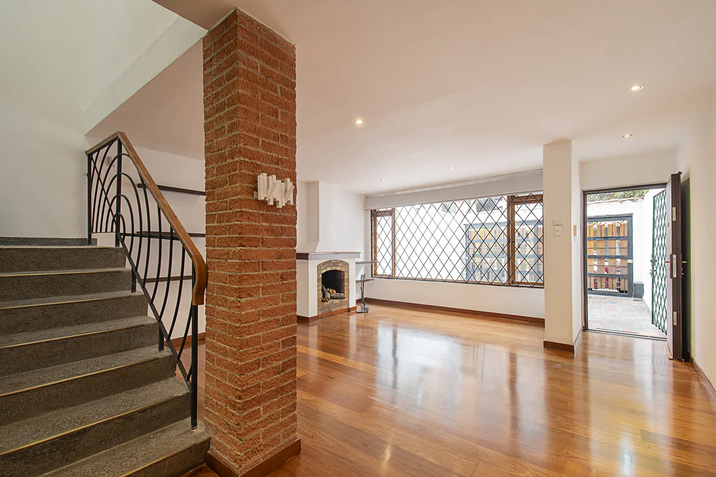 Bright living room with hardwood floors, brick column, fireplace, and large window with diamond pattern. Staircase with black railing on the left.