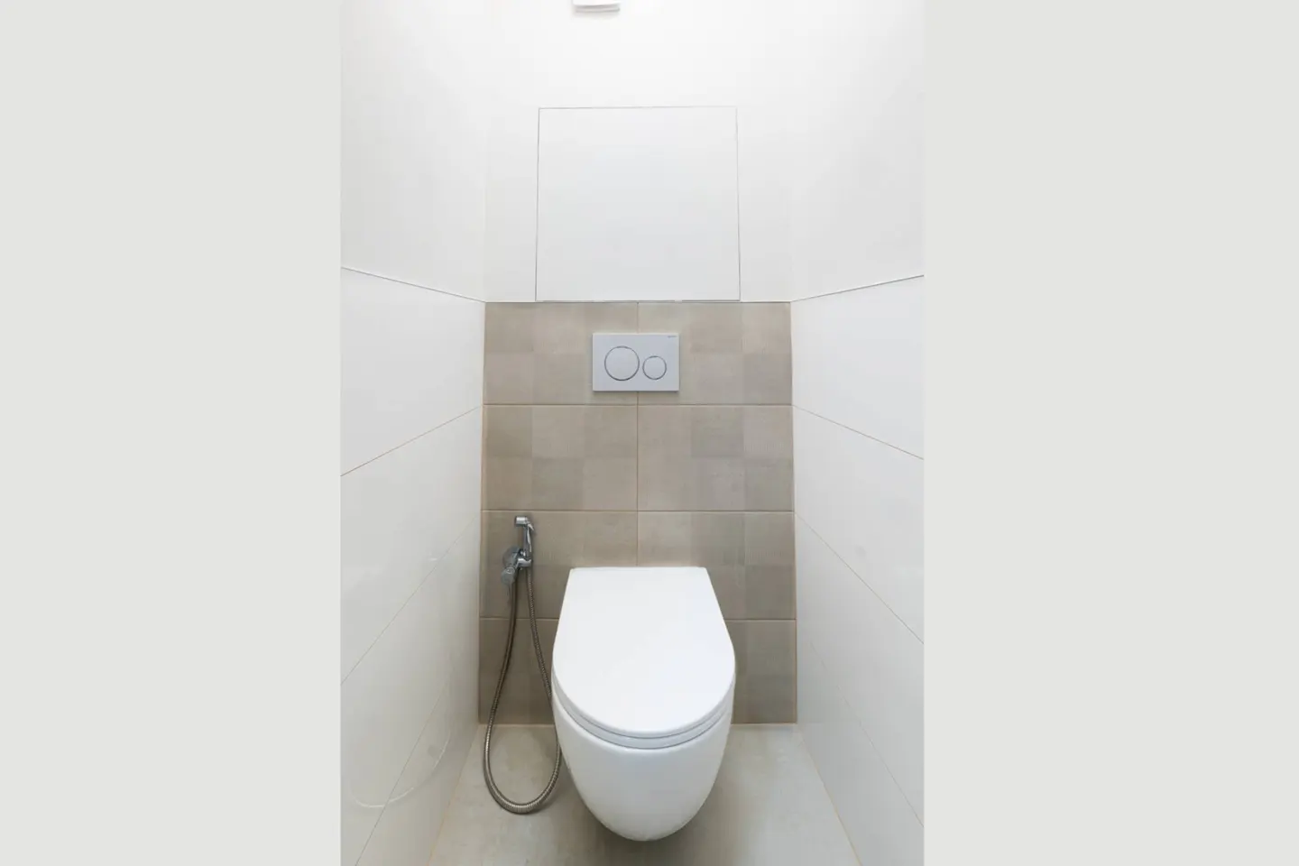 A white toilet with a bidet hose in a small bathroom with white and beige tiles.