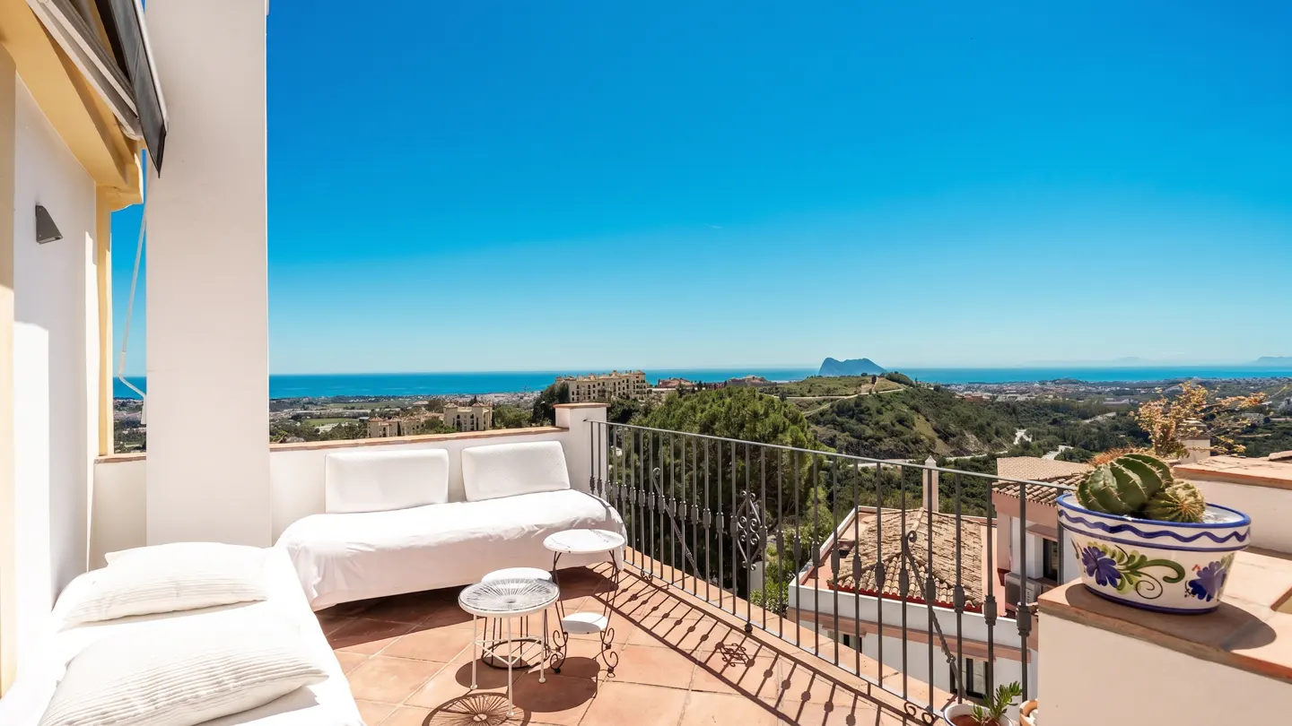 Balcony view with white furniture, terracotta tiles, and wrought iron railing. Distant ocean and blue sky backdrop.