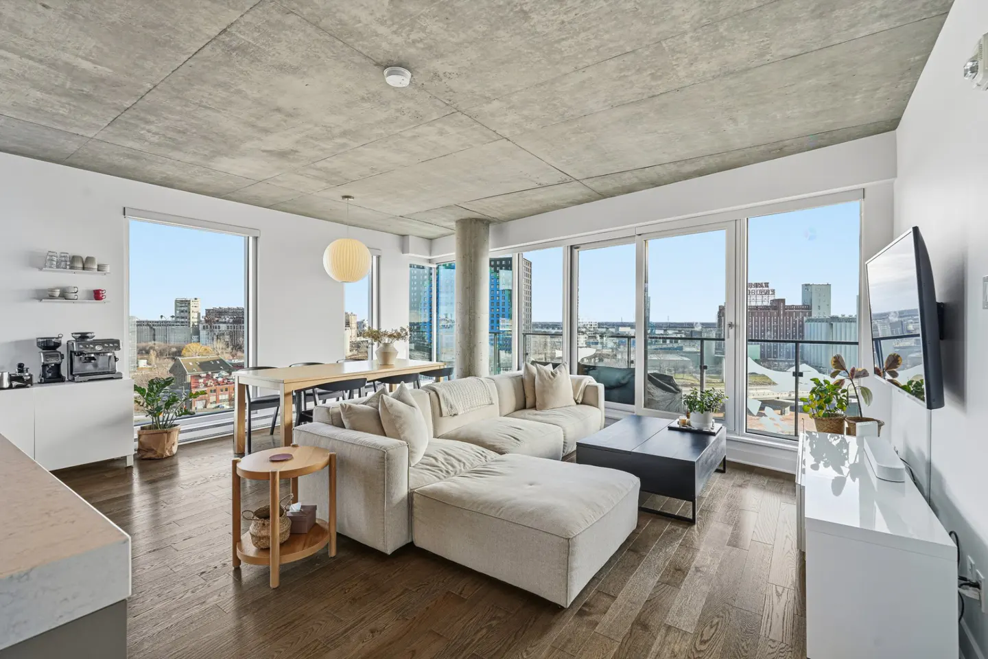 Bright, modern living room with a large white sectional sofa, black coffee table, and floor-to-ceiling windows with a city view.